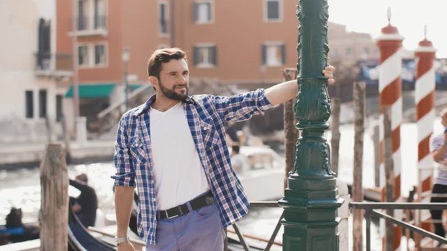 Handsome tourist man standing on the pier against beautiful view on venetian chanal with boats and gondolas in Venice, Italy. Beautiful slow motion 4k footage. Young Caucasian male in casual clothes