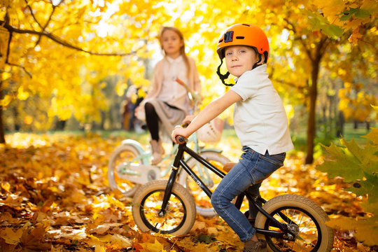 Photo of boy in helmet on running bike, standing girl in autumn park