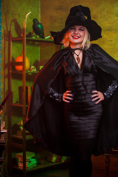 Picture of smiling witch in black hat, dress on background of rack with pumpkin and crow
