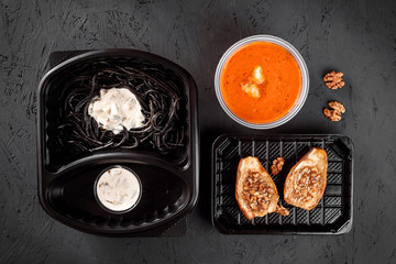 Pasta with cuttlefish ink, tomato cream soup and baked pear with nuts in eco-boxes. on a black background. Healthy food delivery. Vegetarian dish. Vegetarian delivery.