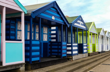 Colourfully painted generic wooden beach huts along a promenade, with steps down to a promenade. Blue sky with white clouds. England.