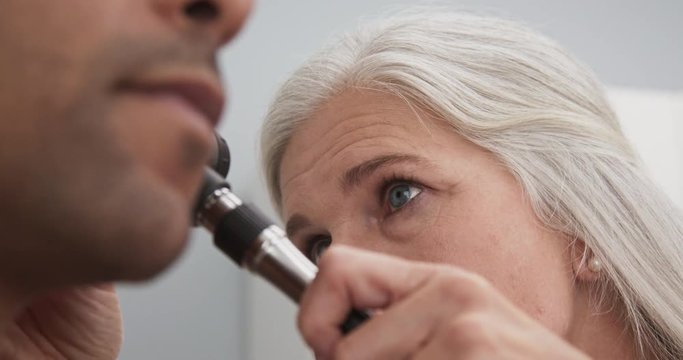 Close up of senior female medical doctor using otoscope to examine patients ear. Young black male patient having doctors appointment for routine checkup