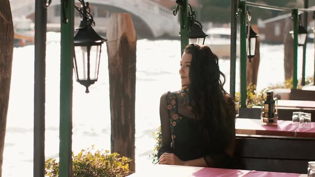 Attractive romantic woman sitting on the pier cafe against beautiful view on venetian chanal with boats and gondolas in Venice, Italy. Slow motion 4k footage. Gorgeous mixed race Asian Caucasian