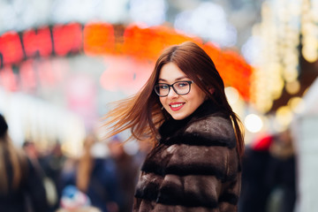 girl in a fur coat and glasses waving her hair and looking at the camera lens against the background of the fair and people