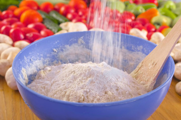 Woman's hands is kneading the yeast dough with a wooden spatula in blue bowl on the table with vegetables.