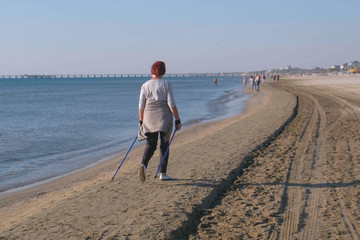 Old woman engaged in Nordic walking, walking on the beach in coastal town. Back view.