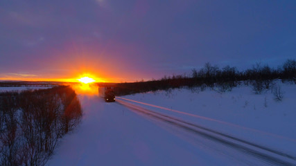 Semi truck driving on snow covered highway trough forest landscape at sunset