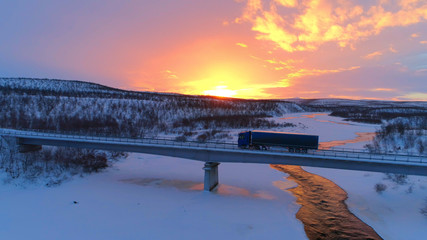 AERIAL: Semi truck crossing the bridge above icy river in the winter at sunset - obrazy, fototapety, plakaty