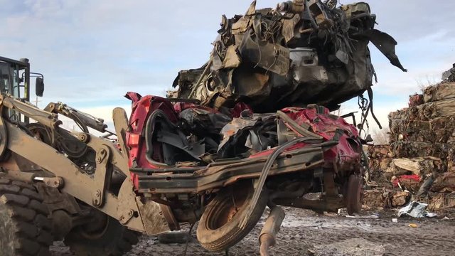 Crushed cars in junk yard are lifted up by forklift and pass by at sunset towards salvage piles in the distance. 