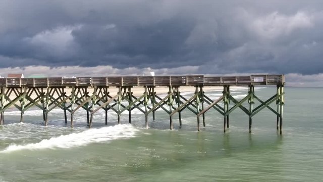 Slow motion shot of ocean waves crashing against a pier off the beach. Dark clouds cover the sky, a storm appears to be brewing.