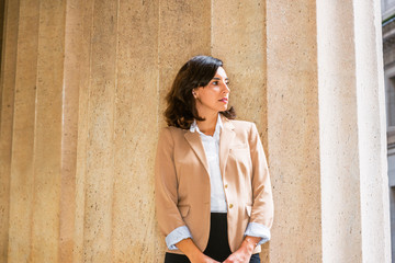 Portrait of Young American Businesswoman in New York City, wearing beige color blazer, unbuttoned, white shirt, black skirt, standing against column outside office building, looking away, thinking..