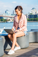 Young American Woman traveling, working in New York, wearing pink leather jacket, blue Denim shorts, white sneakers, sitting by river, working on laptop computer. Brooklyn buildings on far background