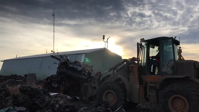 Heavy Machine At Junk Yard Before Sunset Works On Lifting Salvaged Cars While Backing Up To Face Camera. 