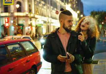 Couple in love on date. Boyfriend holds mobile phone. She hugs him and smokes an electronic...