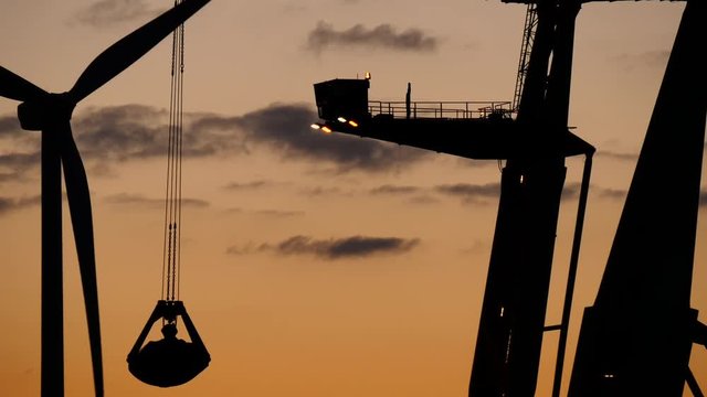 Cargo cranes working in the evening at sundown, discharging a bulk carrier. Windmill is turning around, 4k clip.