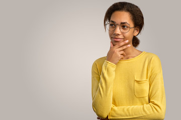 Thoughtful young African American woman holding her chin, smiling, has dark healthy skin, wears a yellow sweater, thinks about something.  Isolated on white background in Studio