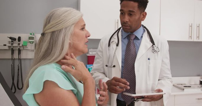 Attractive black millennial doctor using tablet computer while consulting senior patient. Charming elderly woman meeting with young doctor about her neck pain