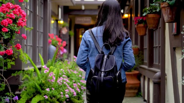 back view of young asian lady walking in the path in carmel ca. little alley full of beautiful flowers and sun shime. elegant woman in jean jacket having fun traveling outdoors during holidays in us.