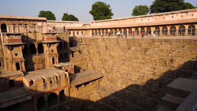 Panning shot of people viewing the beautiful architecture of the chand baori stepwell in Rajasthan. The geometrically made steps and stone palace served as a water storage for the dry areas around the