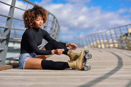Young smiling black girl sitting on urban bridge and puts on skates.