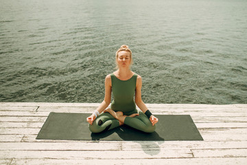 Young attractive woman practicing yoga at lake shore. Lotus asana. Green outfit.Toned.