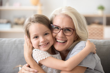 Happy old grandmother hugging little grandchild girl looking at camera, smiling mature mother or senior grandma granny laughing embracing adopted kid granddaughter sitting on couch, headshot portrait - obrazy, fototapety, plakaty