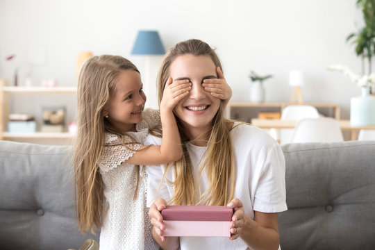 Little cute daughter closing eyes of excited mommy holding gift box sitting on couch, cute preschool child girl making surprise for happy mom, present for mothers day or birthday from kid concept
