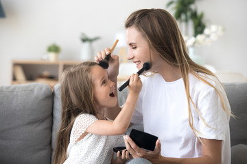 Smiling mom and kid preschool daughter doing makeup together, excited little girl holding make-up brush puts powder on mothers face, happy child applying blush having fun playing with mommy at home