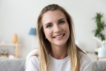 Headshot front portrait of smiling friendly millennial woman sitting alone on couch at home looking...