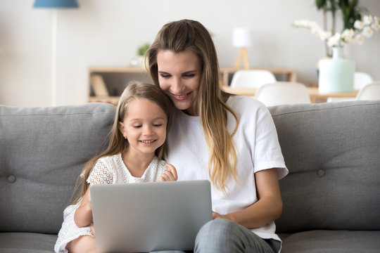 Smiling mother and kid daughter having fun shopping online sitting on sofa, happy mom baby sitter teaching little child girl use laptop application, watching cartoon or making video call on computer