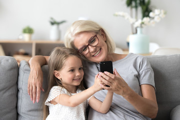 Happy grandmother and cute granddaughter using cellphone together, smiling older grandma and child...