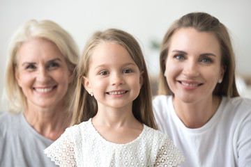 Adorable little girl looking at camera with mom and grandma at background, smiling preschool happy child with beautiful face posing for headshot portrait with mother and grandmother, 3 generations