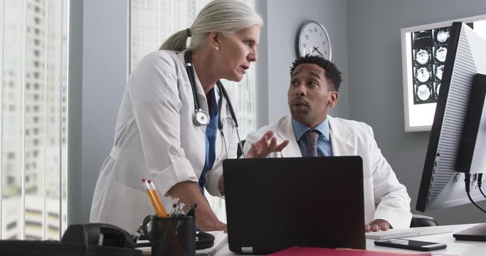Portrait of millennial black doctor using computer while senior colleague directs him. Two doctors working inside medical office looking at computer monitor and typing