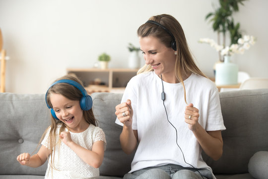 Happy Young Mother And Funny Child Daughter Having Fun Laughing Dancing Sitting On Couch Listening To Music In Earphones Together, Smiling Mom And Kid Wearing Headphones Enjoy Favorite Songs At Home