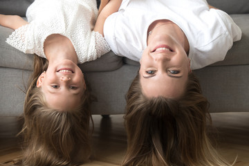 Happy loving mother and little preschool daughter lying upside down on sofa looking at camera, smiling mom and cute funny kid girl having fun, cheerful carefree mommy and child headshot portrait