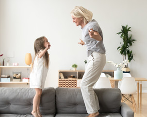 Cute little girl having fun playing with smiling grandmother jumping on couch together, happy...