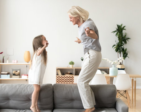 Cute little girl having fun playing with smiling grandmother jumping on couch together, happy granny and active kid grandchild dancing on sofa, grandma and granddaughter laughing playing at home
