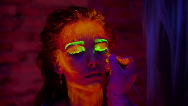 A hand of artist painting a face of beautiful girl with glowing paint with brush. Pretty woman with glowing bodyart in black lamp light.