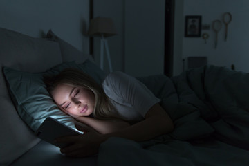 Young sleepy tired woman with half-closed tired eyes lying in bed under the blanket using smartphone at late night, can not sleep.Insomnia, nomophobia, sleep disorder, sleeplessness concept