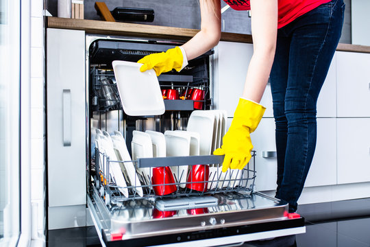 Young woman taking out clean dishes from dishwasher machine.