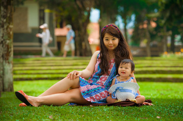 happy and playful Asian Korean woman as loving mother enjoying with sweet and beautiful daughter baby girl sitting together playing on grass at city park in love concept