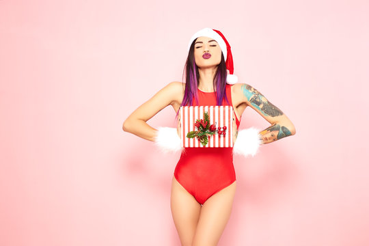 Girl with purple hair tips and tattoo on her arm dressed in red swimsuit, Santa's hat and white fur bracelets holds a christmas gift in her hands on the background of pink wall