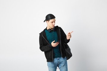 Guy with a black backpack on his shoulder and headphones around his neck dressed in a dark t-shirt, jeans, sweatshirt and a cap uses mobile phone in the studio on a white background