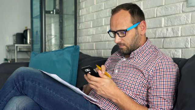 Young man calculating bills on smartphone sitting on sofa