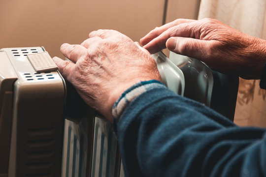 An Elderly Man Warms His Hands Over An Electric Heater. In The Off-season, Central Heating Is Delayed, So People Have To Buy Additional Heaters To Keep Houses Warm Despite Increased Electricity Bills