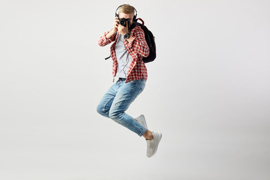 Blond guy in headphones, with black backpack on his shoulder dressed in a white t-shirt, red checkered shirt and jeans jumps and makes photos on camera on the white background in the studio