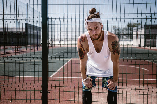 Young man with white headband  on his head and tattoos on his arms dressed in the white t-shirt, black leggings and blue shorts stands next to the playground fence outside on a sunny day