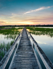 coastal waters with a very long wooden boardwalk pier in the center during a colorful summer sunset under an expressive sky with reflections in the water and marsh grass in the foreground