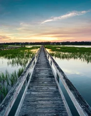 Foto auf Acrylglas coastal waters with a very long wooden boardwalk pier in the center during a colorful summer sunset under an expressive sky with reflections in the water and marsh grass in the foreground © makasana photo