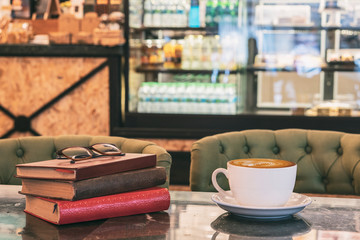 Cappuccino coffee in a white cup on a wooden table with a modern coffeeshop in the background and books, glass, selective focus.